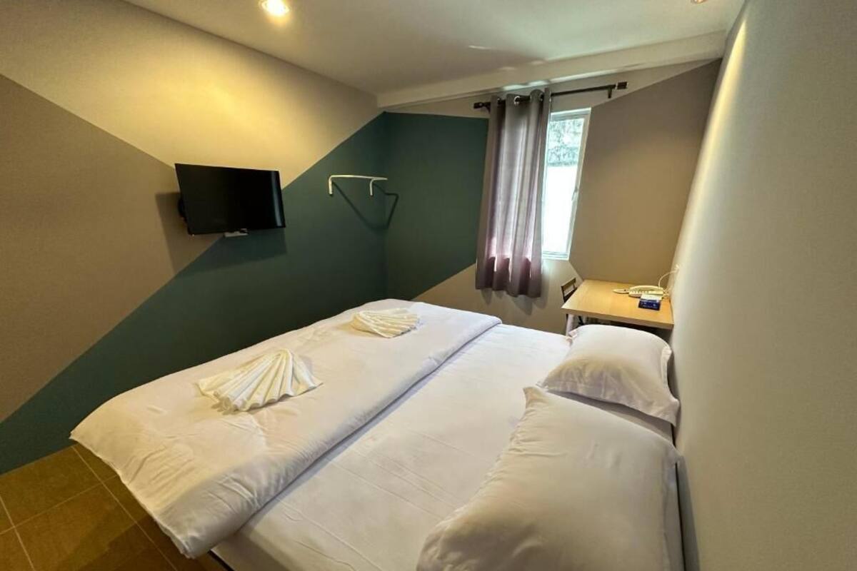 Ett hotellrum med två sängar, en platt-TV som är monterad på väggen, ett litet bord med en lampa och ett fönster med gardiner.