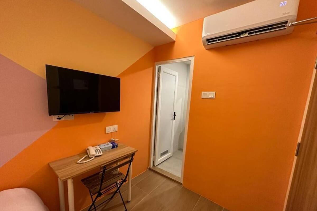 Ett litet rum med orange väggar, en platt-TV som är monterad på väggen, ett skrivbord i trä med en telefon och en luftkonditioneringsanläggning.