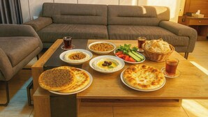 Una mesa de comedor con diversos platos y bebidas, un sofá y una mesita de noche de madera en una habitación con pared de color claro y una ventana.