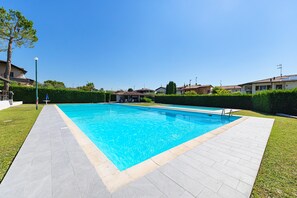 Une piscine rectangulaire entourée d’une terrasse carrelée, située dans un espace aménagé avec de la pelouse et des haies.
