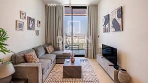 A modern living room with a grey sofa, a coffee table, and a flat-screen TV. There are curtains, framed pictures on the walls, and a large window with a view. - Cozy 1-bedroom apartment in Dubai (Dubai)