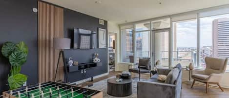A modern living room with a foosball table, a grey sofa, and a large window offering a city view.