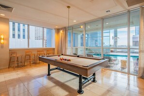 Une table de billard dans une pièce dotée de grandes fenêtres, avec vue sur une piscine et sur le paysage urbain.