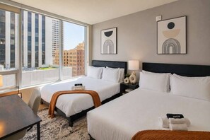 Une chambre d’hôtel avec deux lits, un bureau et une vue sur les bâtiments, visible à travers de grandes fenêtres.