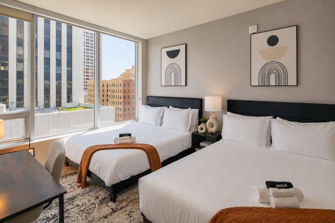 A hotel room with two beds, a desk, and a view of buildings through large windows.