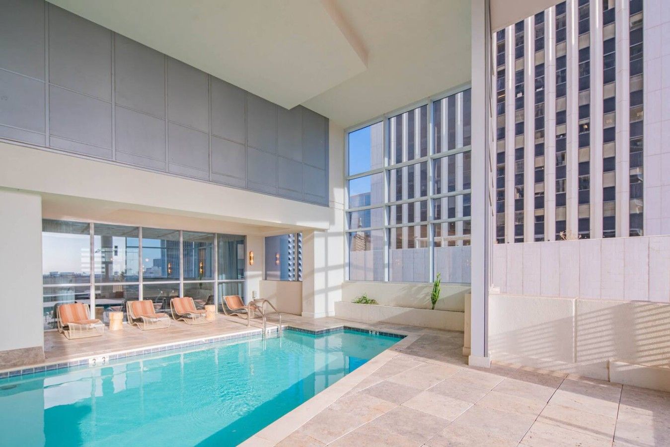 An indoor swimming pool with lounge chairs and a view of a cityscape through large windows.