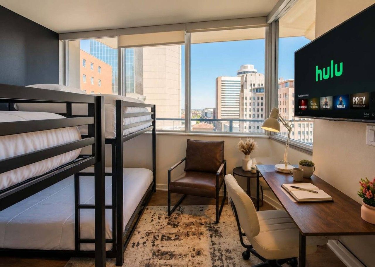A room with bunk beds, a desk with a lamp, a chair, a potted plant, and a TV displaying the Hulu logo.