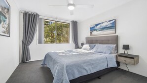 A bedroom with a bed, a nightstand, a ceiling fan, a window with curtains, and a painting on the wall. - Flynns Beach Seascape (Port Macquarie)