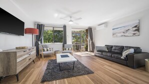A modern living room with a wooden floor, a grey sofa, a white coffee table, and a TV mounted on the wall. - Flynns Beach Seascape (Port Macquarie)