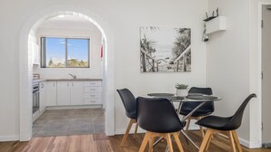 A modern kitchen with white cabinets and a window, a dining area with a round table and black chairs, and a beach-themed painting on the wall. - Flynns Beach Seascape (Port Macquarie)