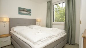 Ein ordentlich bezogenes Bett mit weißen Leinen, zwei Nachttische mit Lampen, ein Fischgemälde an der Wand und ein Fenster mit Vorhängen.