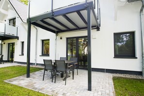 Een terras buiten met een overdekte tafel en stoelen, een wit gebouw met grote ramen en een verhard gebied.