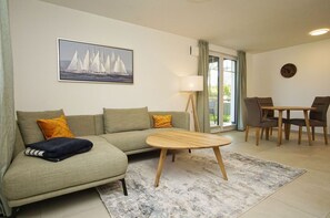 A living room with a sofa, a coffee table, a dining table with chairs, a lamp, and a painting of sailboats. - Beachfront apartment with terrace - Rex Rugia House 17 Apartment 04 - Beach Resort (Mönchgut)