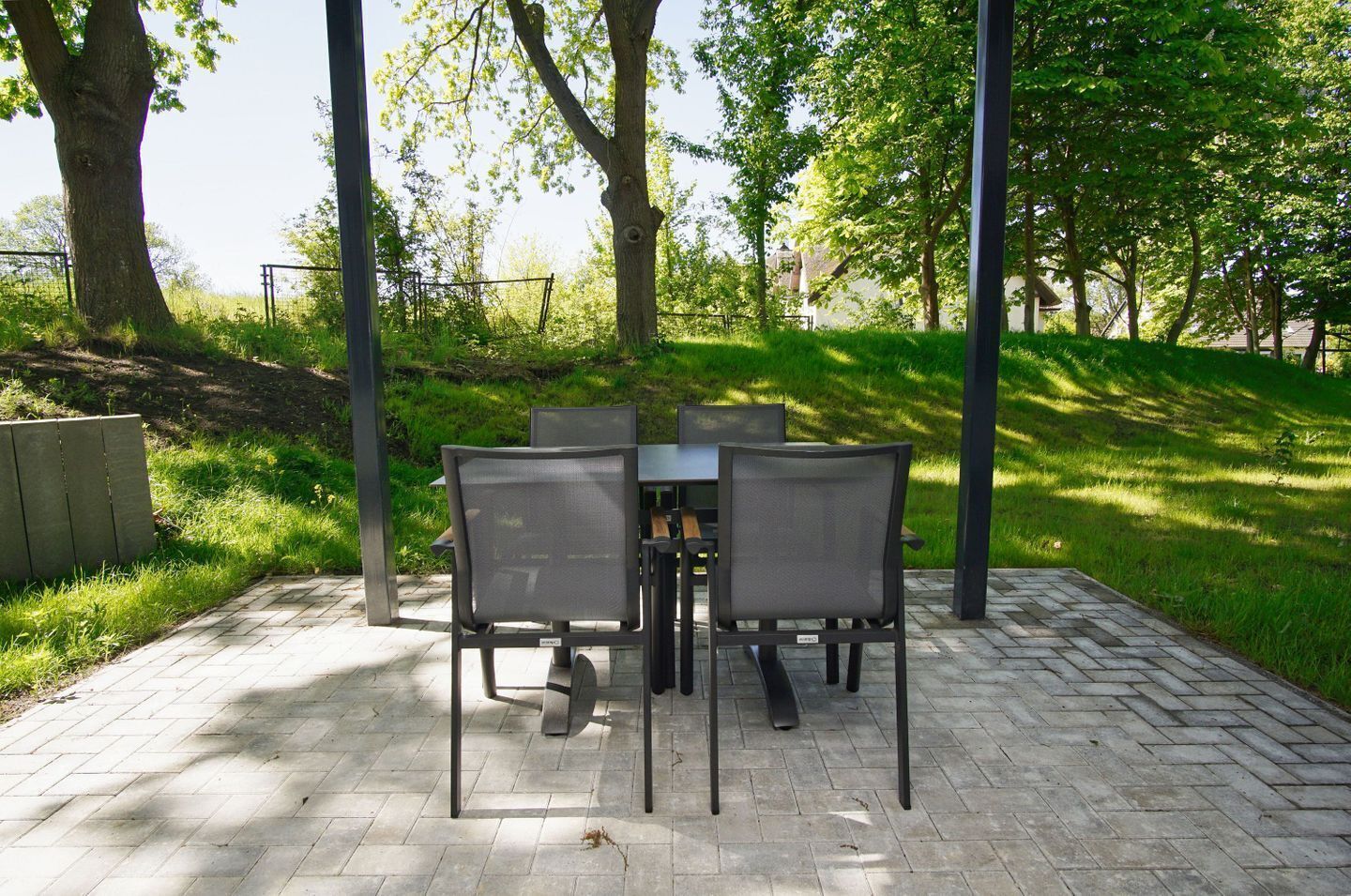 Een terras buiten met een tafel en stoelen, omgeven door bomen en een met bakstenen bestraat pad.