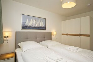 A hotel room with a bed, two bedside lamps, a painting of sailboats, and a wardrobe. - Beachfront apartment with terrace - Rex Rugia House 17 Apartment 03 - Beach Resort (Mönchgut)