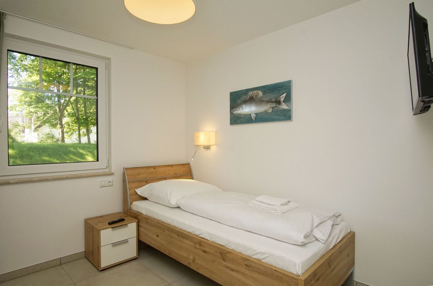 A bedroom with a wooden bed, a nightstand, a drawer, a window with a view of trees, a wall-mounted TV, and a painting of a fish.