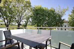 Eine Terrassengruppe mit Tisch und Stühlen, umgeben von Glasgeländern, mit Bäumen und einem Gebäude im Hintergrund.