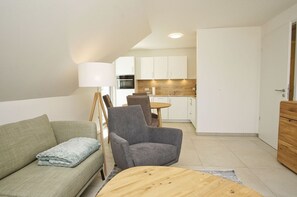 A modern living room with a sofa, armchair, and a wooden coffee table. A dining area with a table and chairs is visible in the background. - Beachfront apartment with balcony - Rex Rugia House 17 Apartment 08 - Beach resort (Mönchgut)