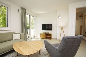 Ein modernes Wohnzimmer mit einer Couch, einem runden Holztisch, einem grauen Sessel, einem an der Wand befestigten Fernseher und einer Holzkommode mit dem Schriftzug "STRAND".