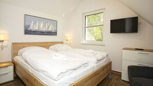 Un dormitorio con cama de madera, ropa de cama blanca, un cuadro de un velero, televisor y ventana con vistas a árboles.