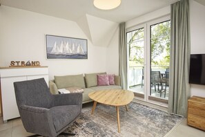 Ein modernes Wohnzimmer mit Sofa, Sessel, Couchtisch und Fernseher. An der Wand hängt ein Gemälde und es gibt ein Fenster mit Vorhängen.