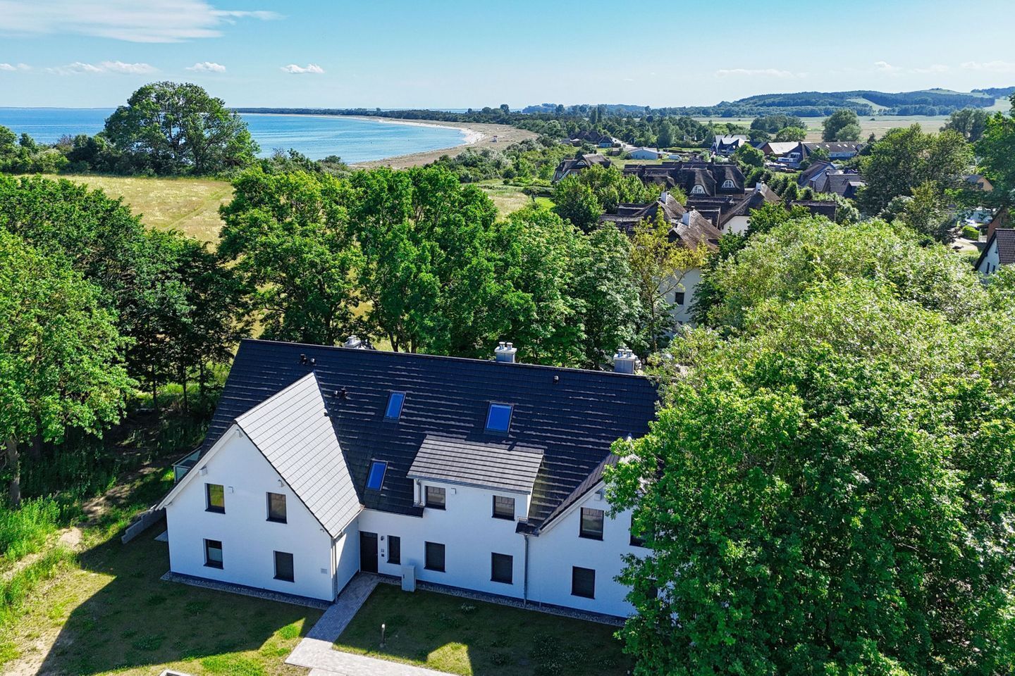 Ein modernes, zweistöckiges Haus mit geneigtem Dach, umgeben von üppigem Grün und Blick auf einen Strand und sanfte Hügel.
