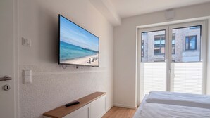 A bedroom with a bed, a TV mounted on the wall, a wooden bench, and a window with blinds. - 2120 - Gode Ström Quartiere "Twee" - Beach meadow - 2120 - Gode Ström Quartiere (Dahme)
