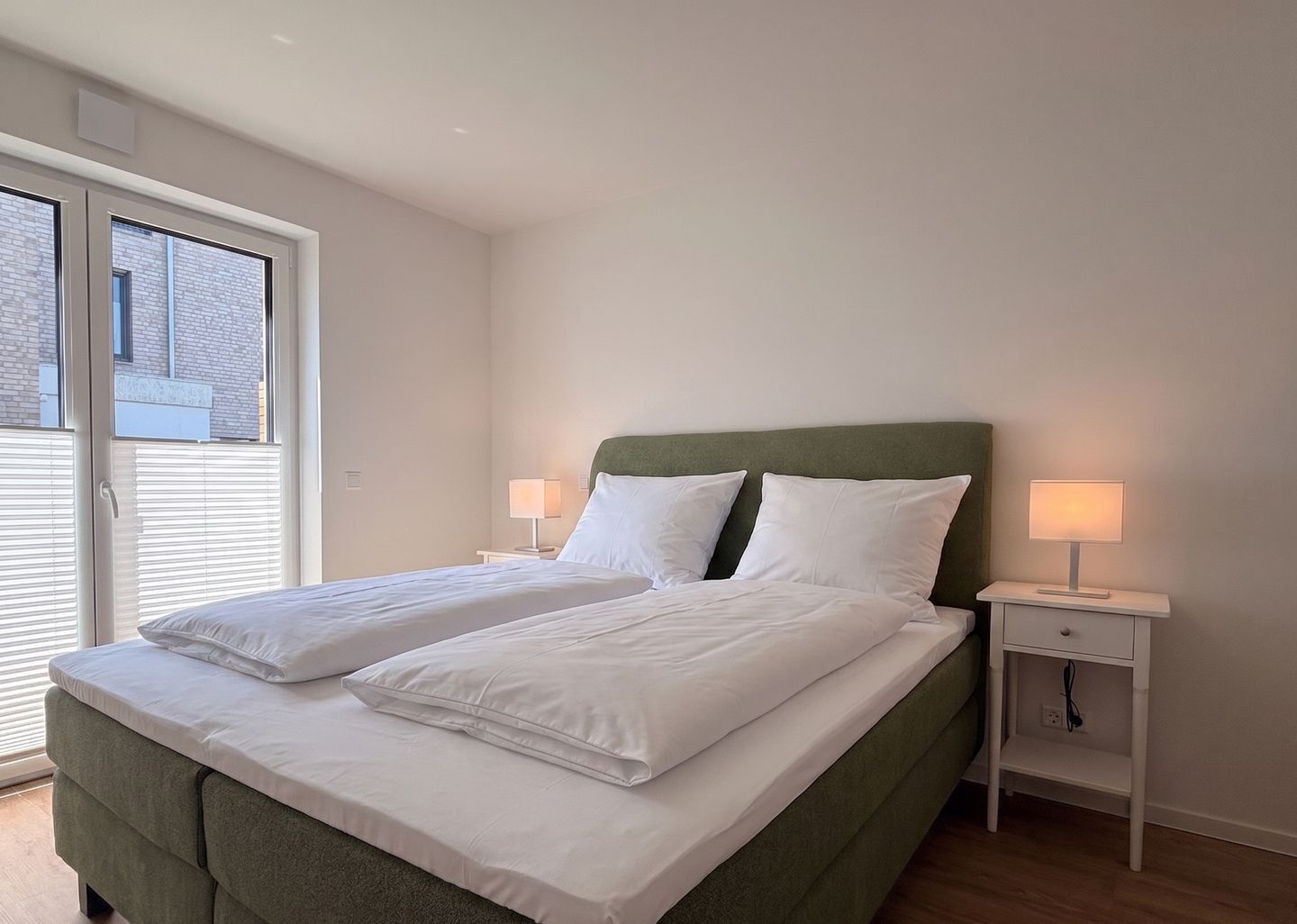 A double bed with white linens, a green upholstered headboard, two bedside tables with lamps, and a sliding glass door to the outside.