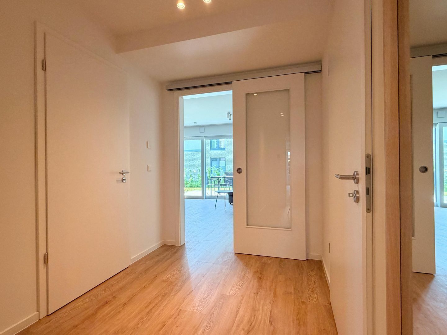 Un pasillo con suelo de madera, paredes blancas y un ventanal corredera que da a un área exterior con una mesa y sillas.