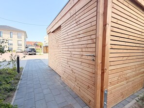 A wooden building with a closed shutter, a paved walkway, and a parked car in the background. - 2130 - Gode Ström Quartiere "Dree" - Beach meadow - 2130 - Gode Ström Quartiere (Dahme)