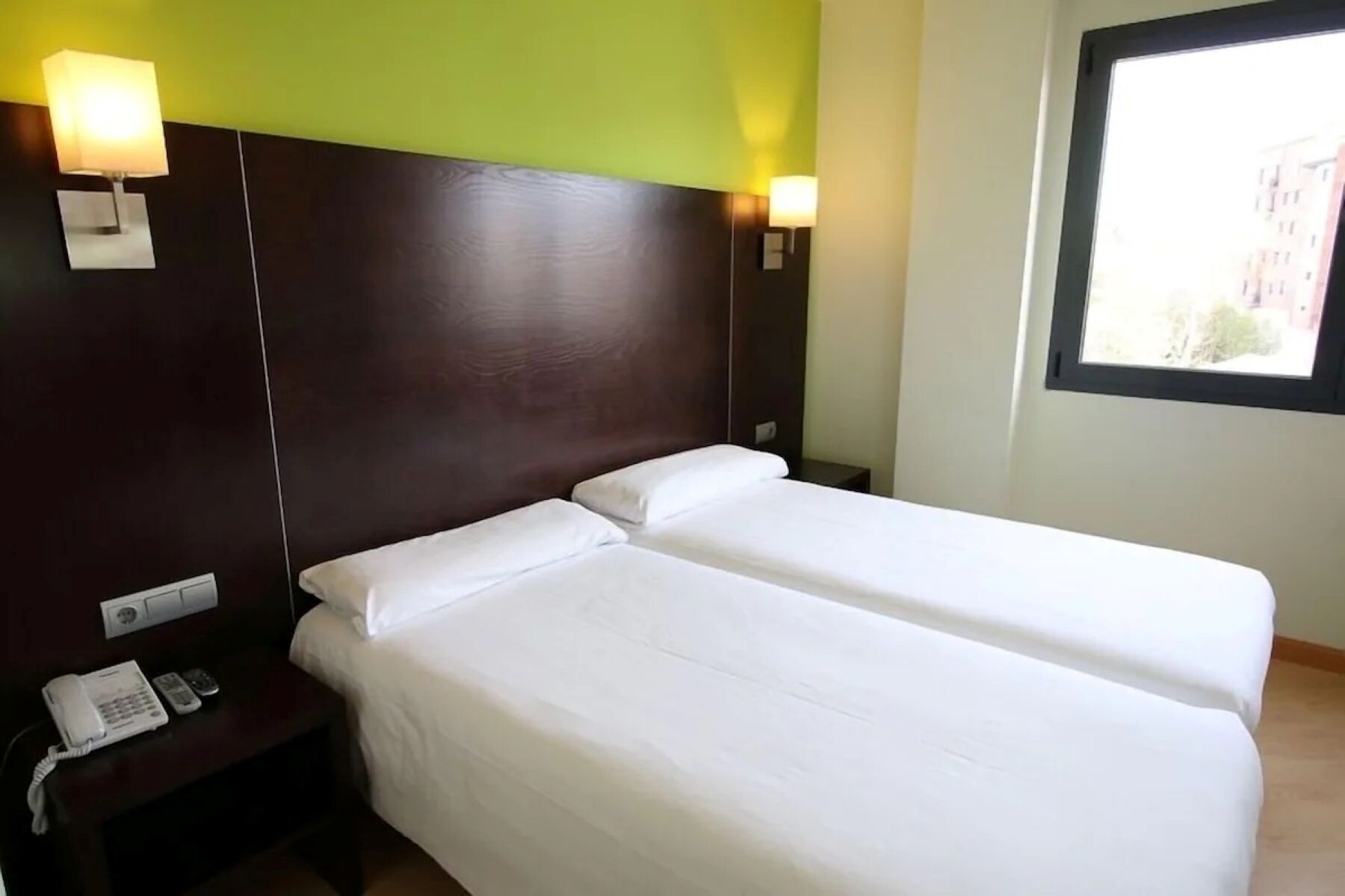 Habitación de hotel con dos camas individuales, teléfono en la mesita de noche y ventana con vistas a edificios.