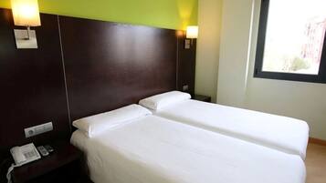 Habitación de hotel con dos camas individuales, teléfono en la mesita de noche y ventana con vistas a edificios.
