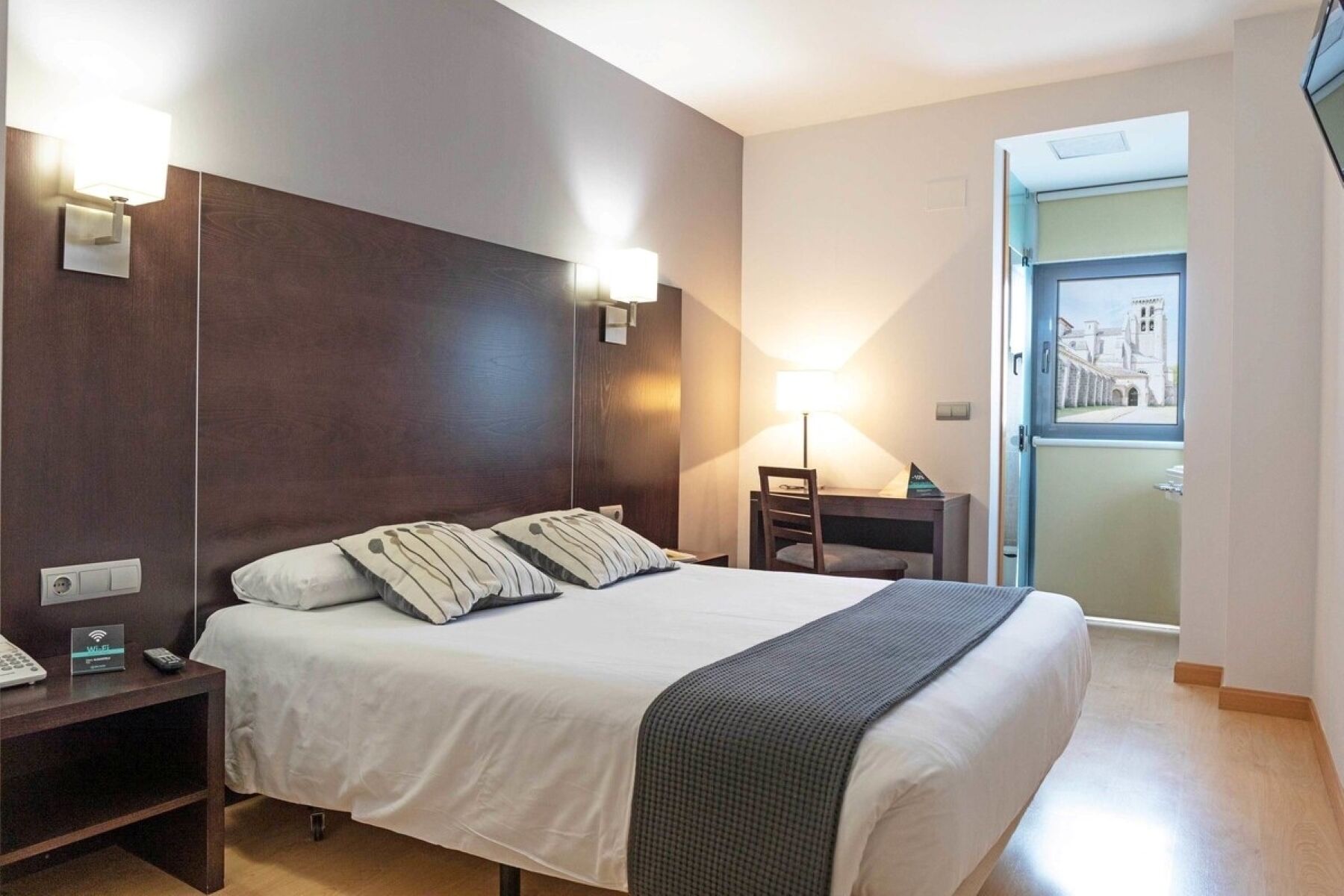 Habitación de hotel con una cama grande, lámparas de noche, un escritorio y vista a un edificio desde la puerta.