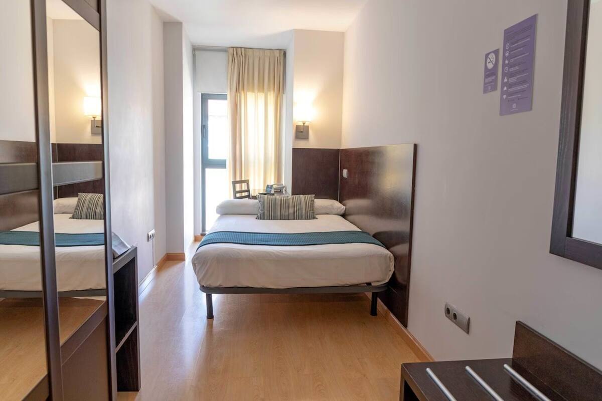 A hotel room with a bed, bedside table, lamp, and a view of the outside through a window with curtains.