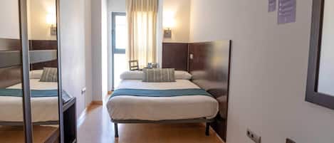 A hotel room with a bed, bedside table, lamp, and a view of the outside through a window with curtains.