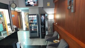 A lobby with a reception desk, a clock, a television, vending machines, and two grey armchairs.