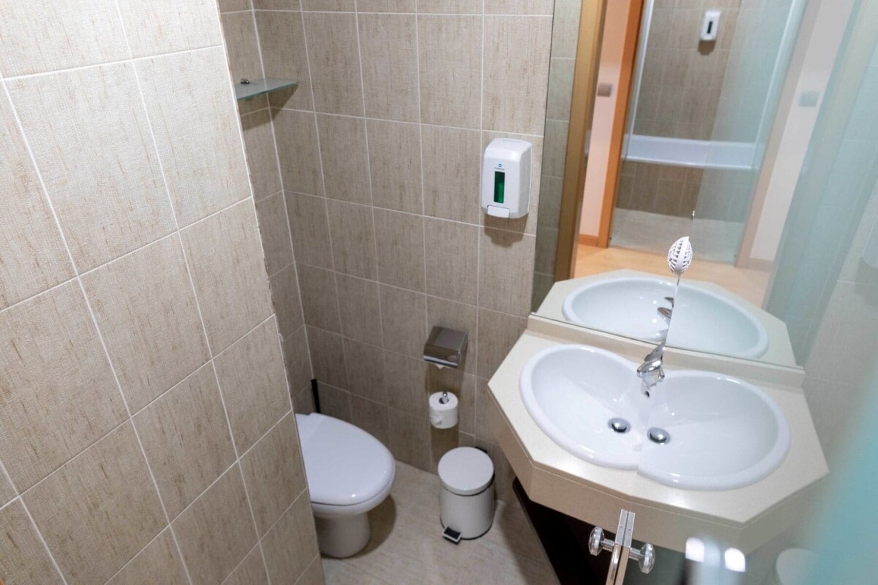 A bathroom with two sinks, a toilet, a trash bin, and a soap dispenser.