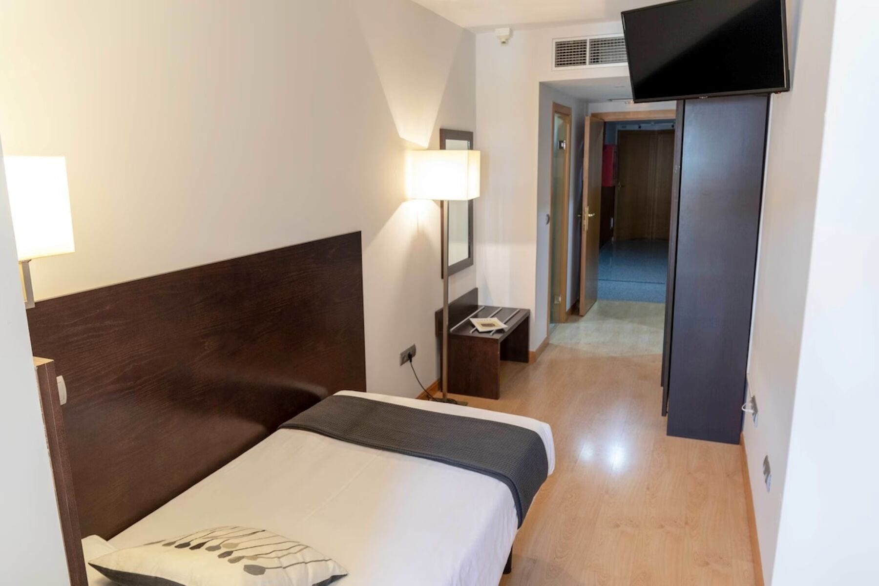 A hotel room with a bed, bedside lamp, desk with a chair, and a television on the wall.