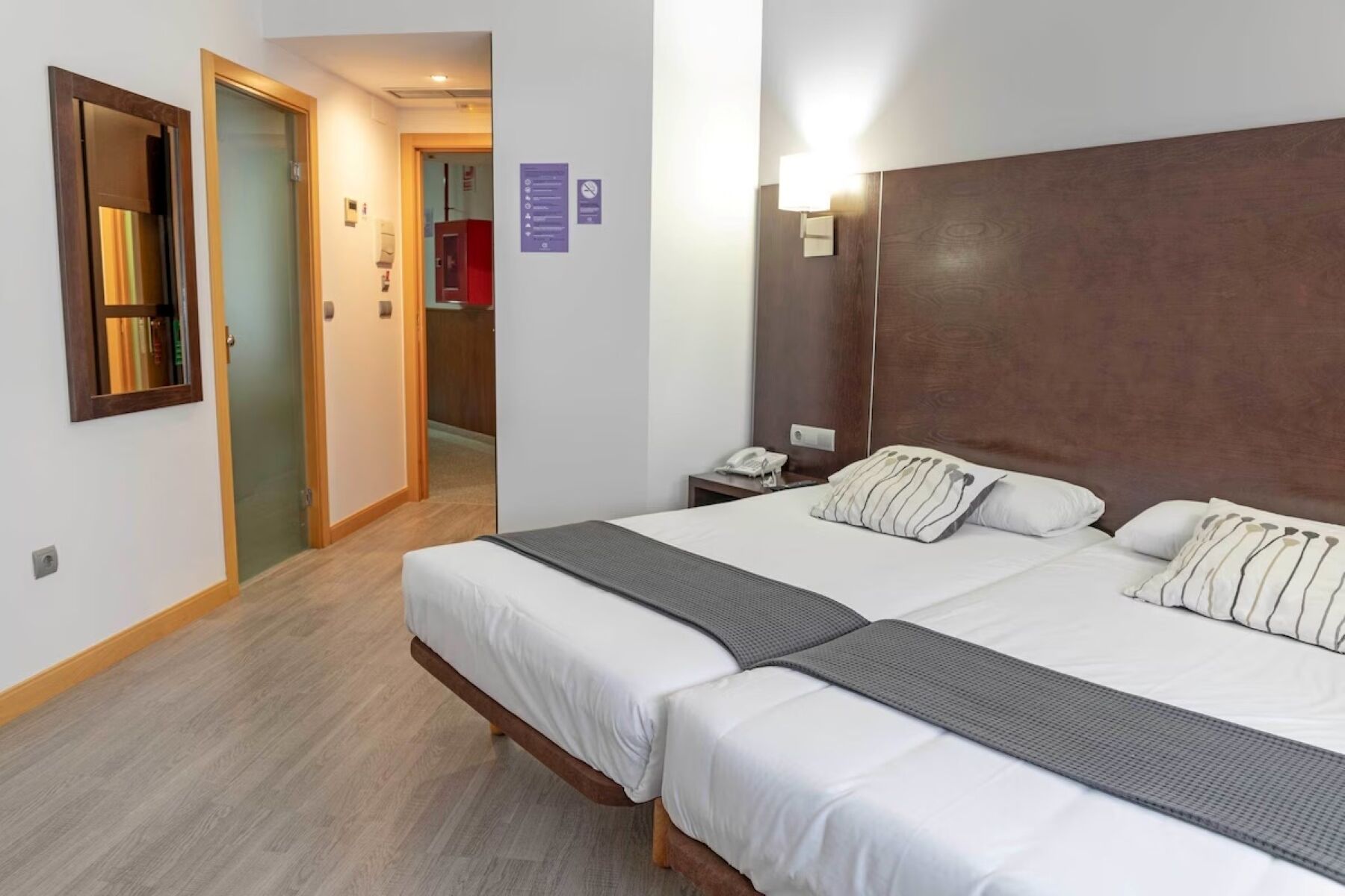 A hotel room with two beds, a bedside table with a phone, and a wall-mounted light.