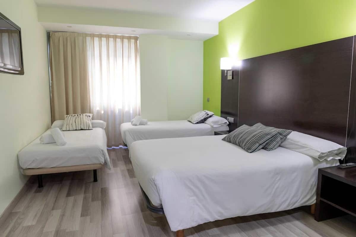A hotel room with two single beds and one double bed, a wooden floor, a green accent wall, and a nightstand with a lamp.