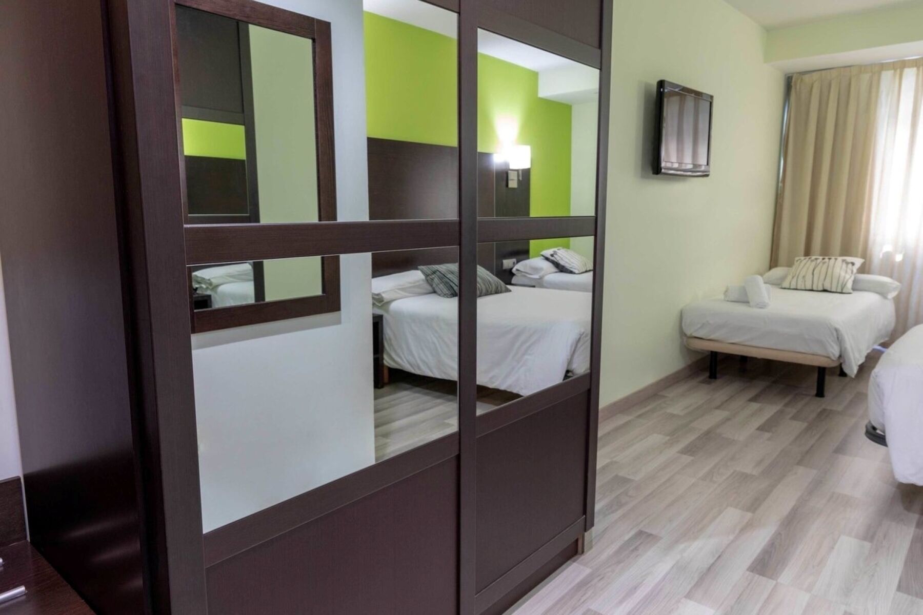 A hotel room with two beds, a mirror, a television, and a window with curtains.