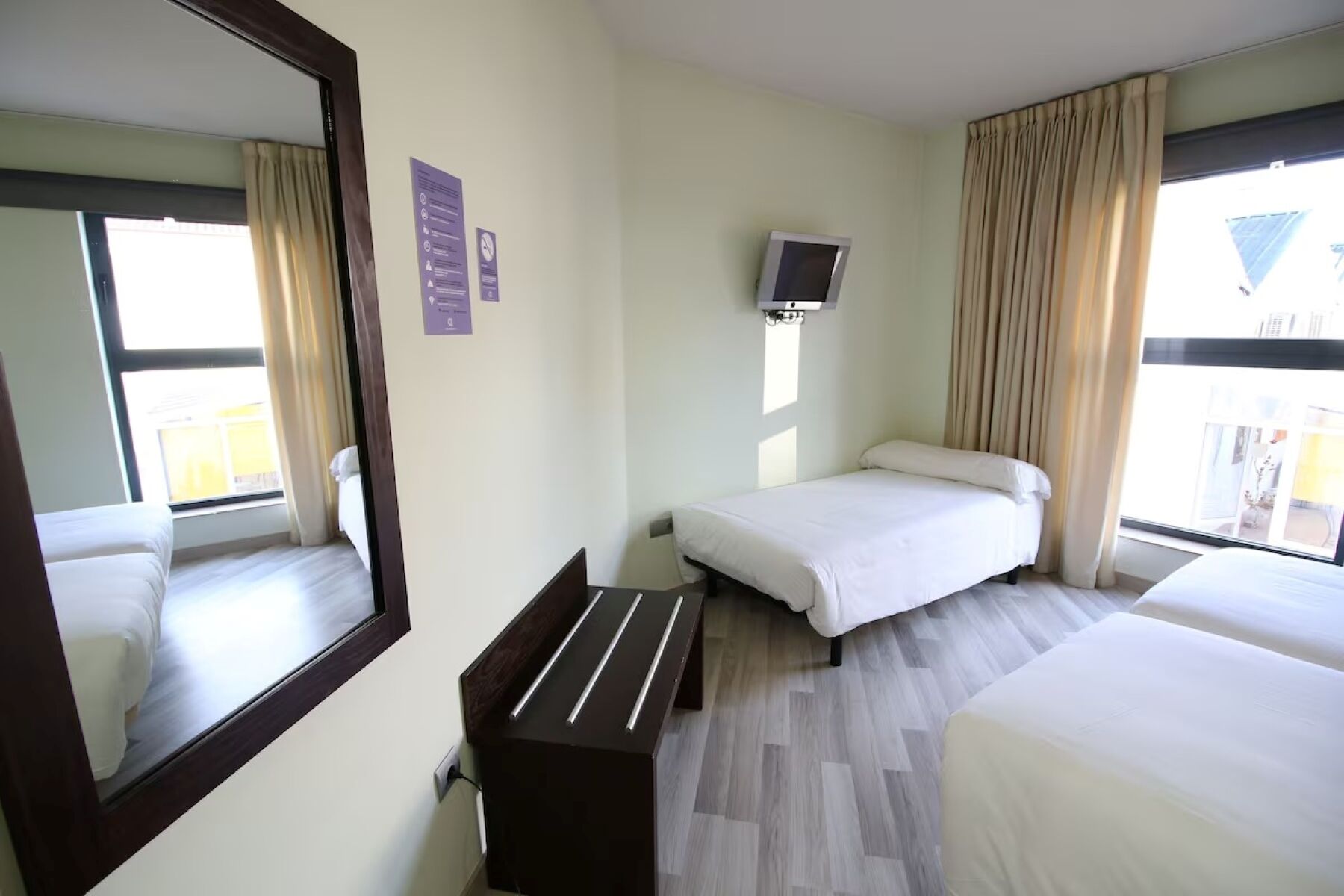 Hotellihuone, jossa on kaksi sänkyä, suuri peili, seinälle asennettu taulutelevisio ja pieni puinen penkki.