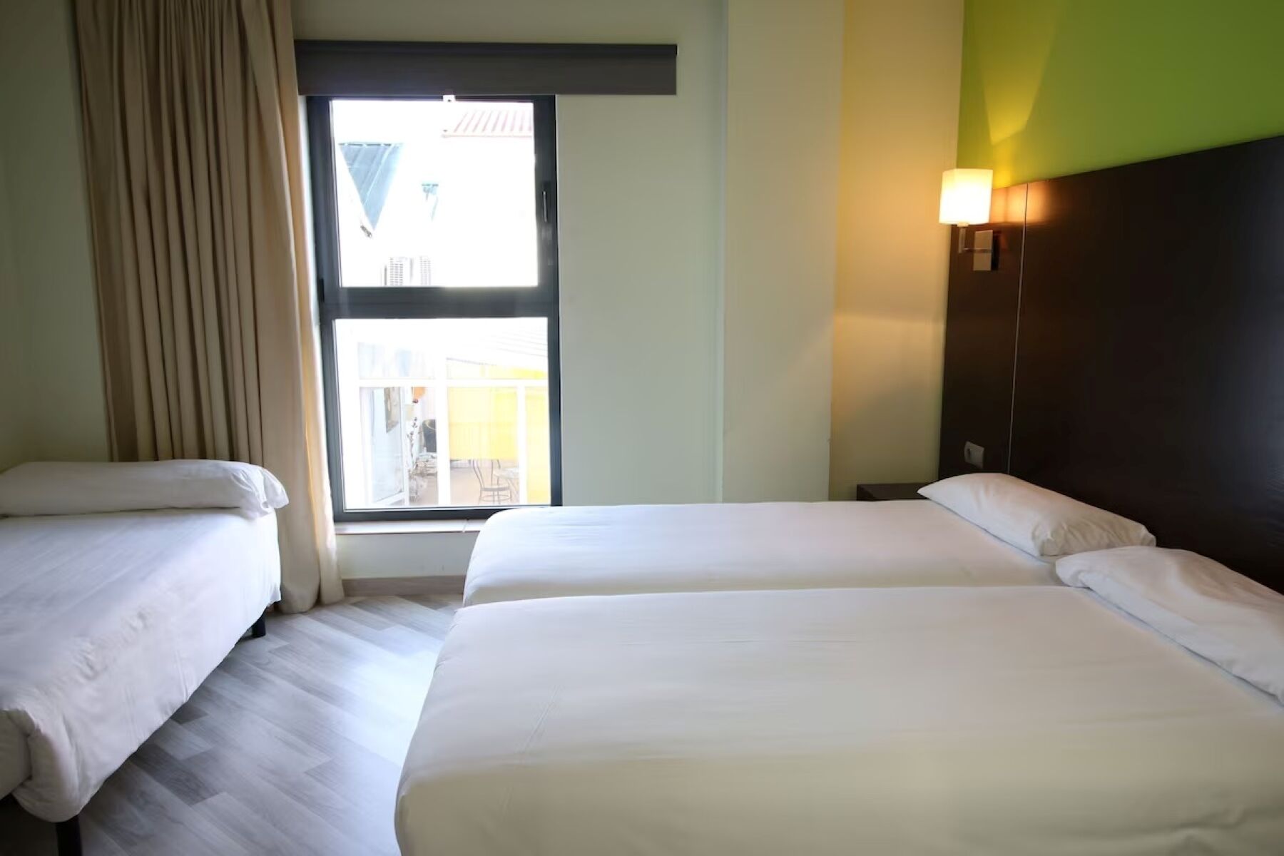 A hotel room with two beds, a window with curtains, a wall-mounted lamp, and a view of the outside.