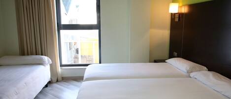A hotel room with two beds, a window with curtains, a wall-mounted lamp, and a view of the outside.