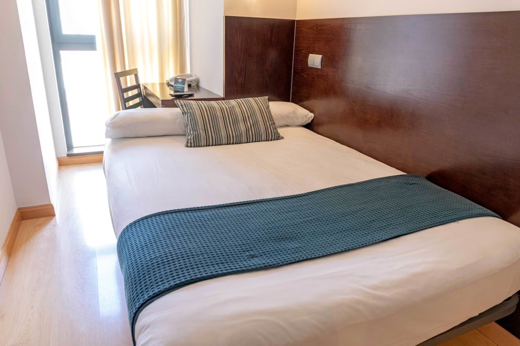 A hotel room with a bed, a wooden headboard, a bedside table with a lamp, and a window with curtains.