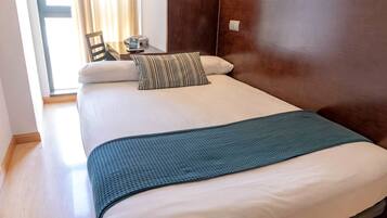 Habitación de hotel con cama, cabecera de madera, mesita de noche con lámpara y ventana con cortinas.