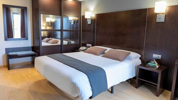 A modern hotel room with a large bed, wooden headboard, bedside tables, a mirror, and a small desk with a lamp.