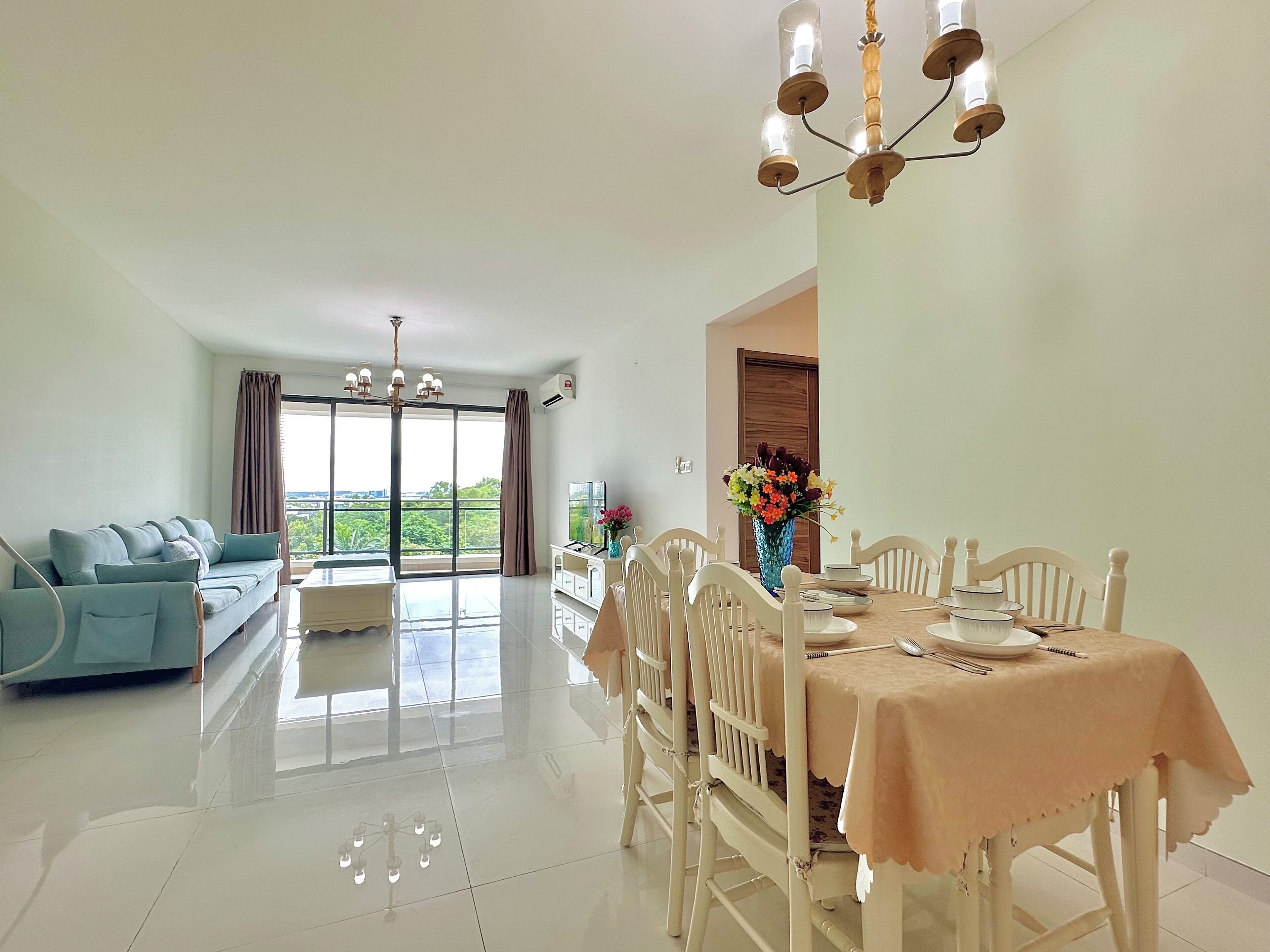 A spacious living area with a sofa, dining table, and chairs. There is a large window providing a view of greenery.