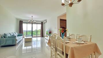A spacious living area with a sofa, dining table, and chairs. There is a large window providing a view of greenery.