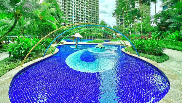 Una piscina con diseño de mosaico, rodeada de exuberante vegetación y altos edificios residenciales.
